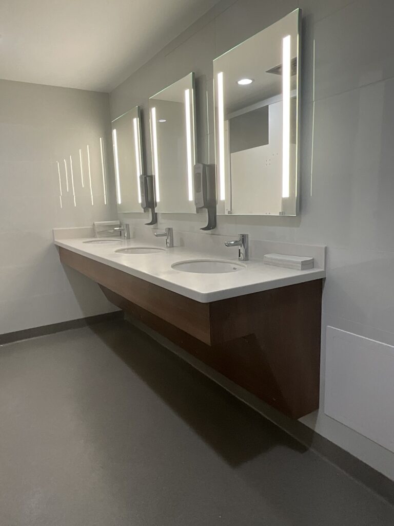 Modern bathroom with a double sink vanity, three illuminated mirrors, and wall-mounted soap dispensers, highlighting contemporary design and functionality.