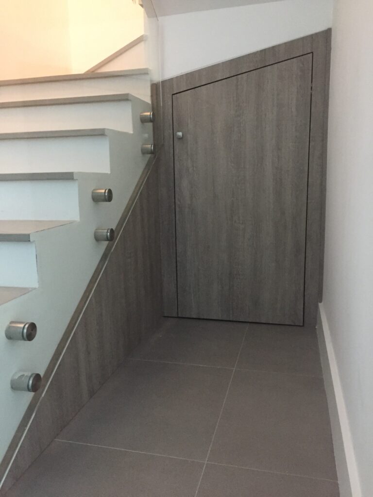A modern staircase with sleek, white steps and stainless steel railings, featuring a hidden door under the stairs within a gray wood-accented wall.
